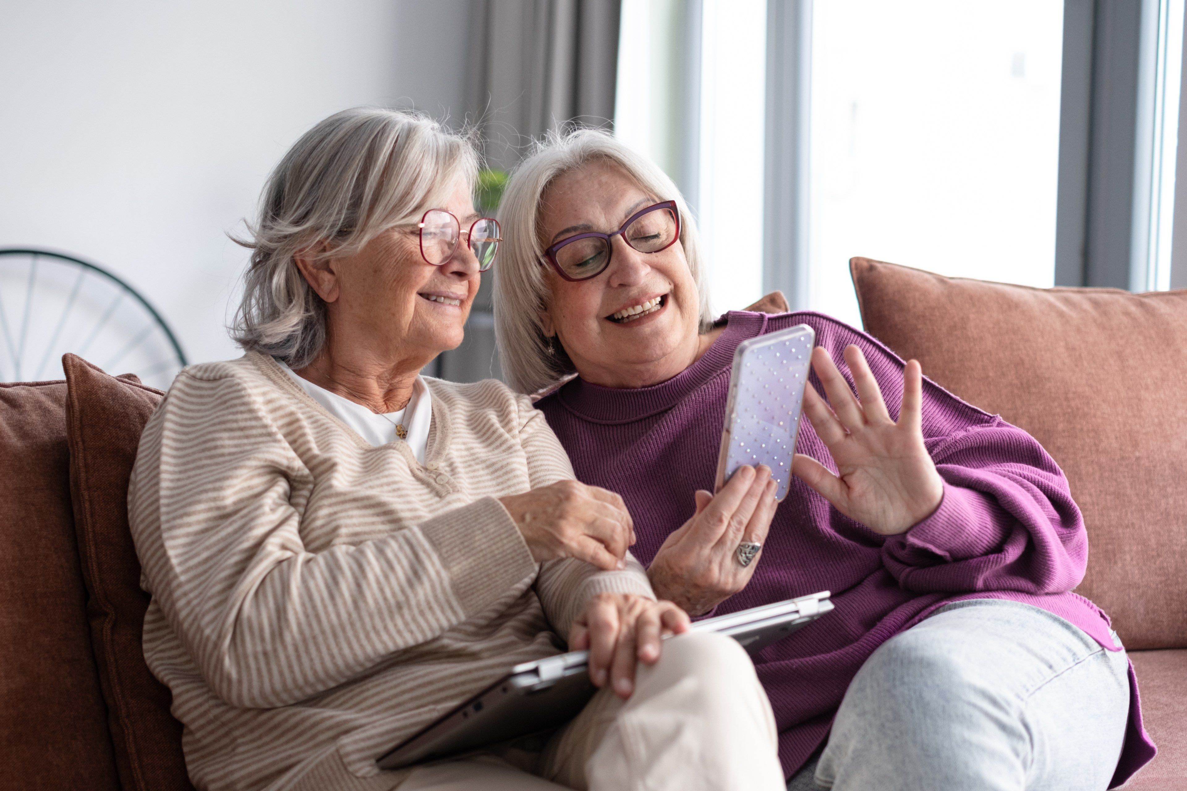 To eldre venninner ler sammen i sofaen og ser på en mobil